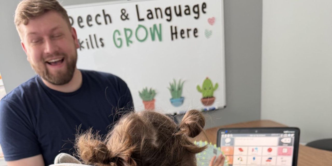 Speech-language pathologist Justin smiles with a young client seated in front of a communication device during a speech therapy session