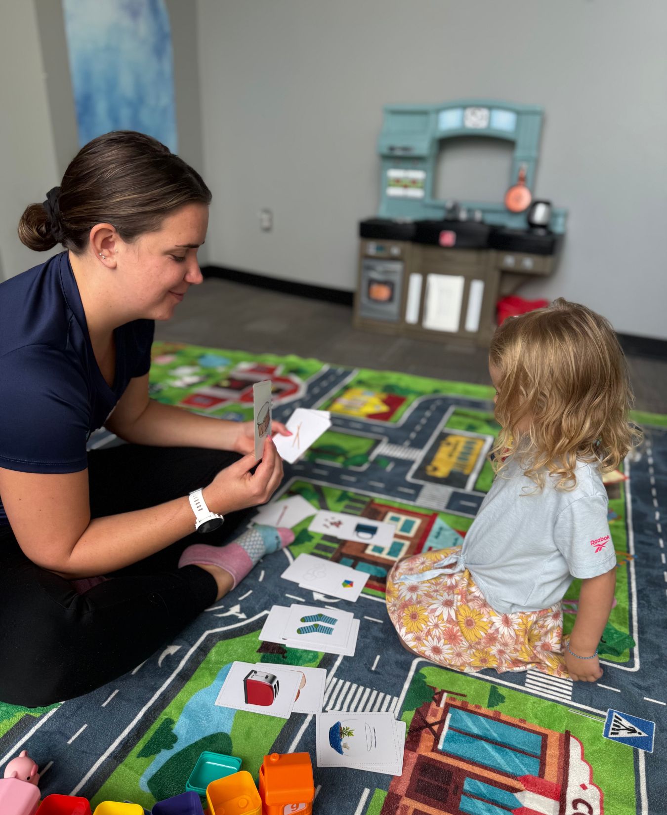 A speech-language pathologist holds up a picture card for a toddler sitting on a play mat during a speech therapy session at Empowerize Therapy Center