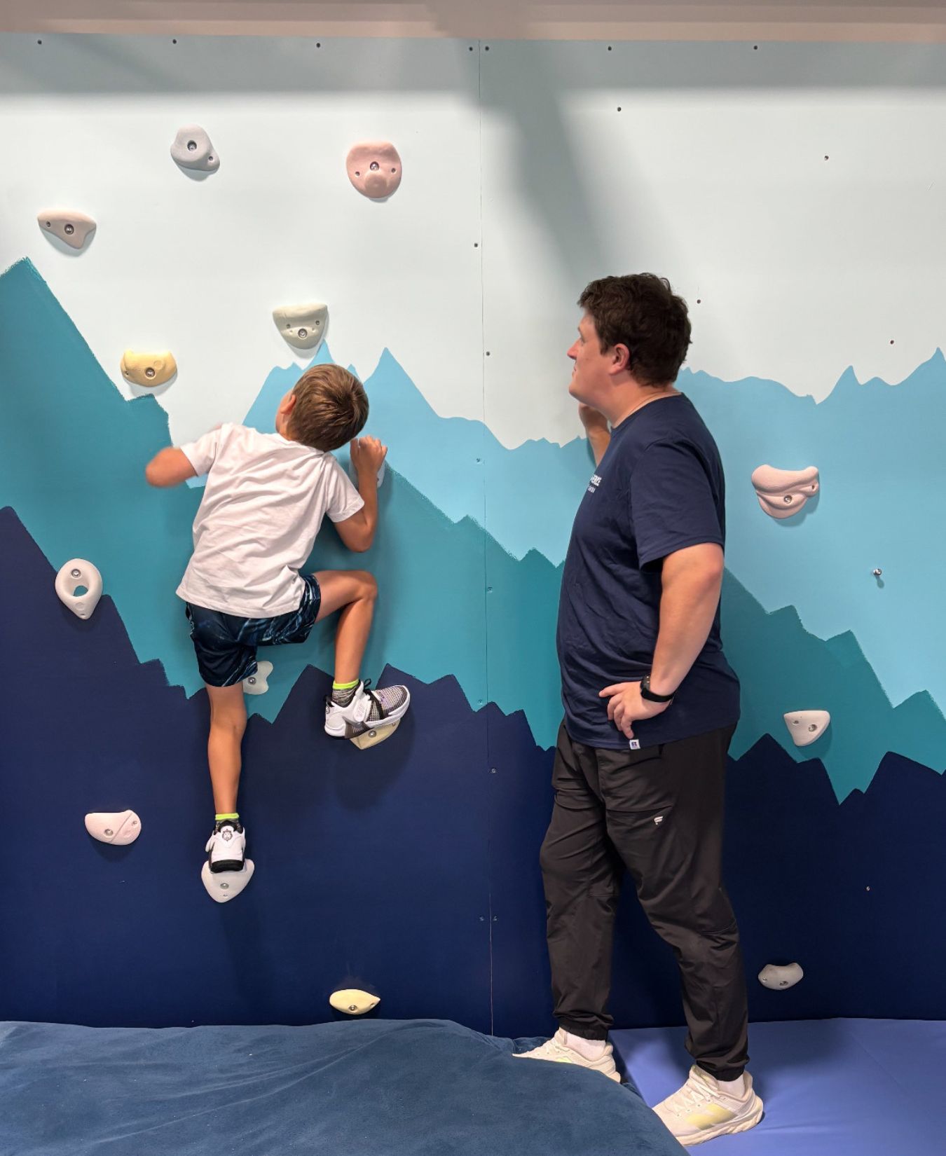 A child climbs the therapy wall while a therapist spots him